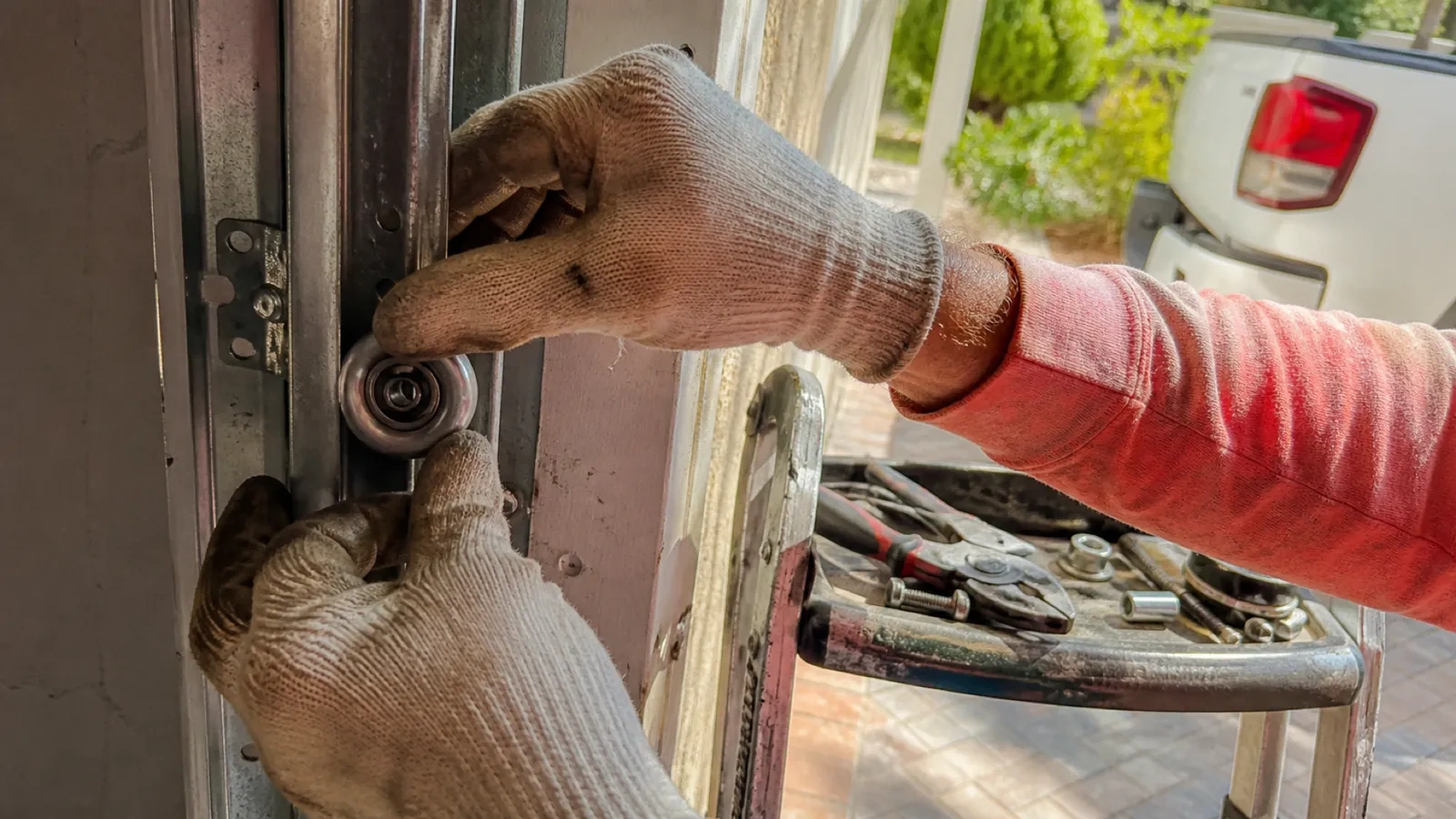 Gloved hands holding a metal roller wheel against a door track, with pliers and small hardware on a stepladder tray nearby.
